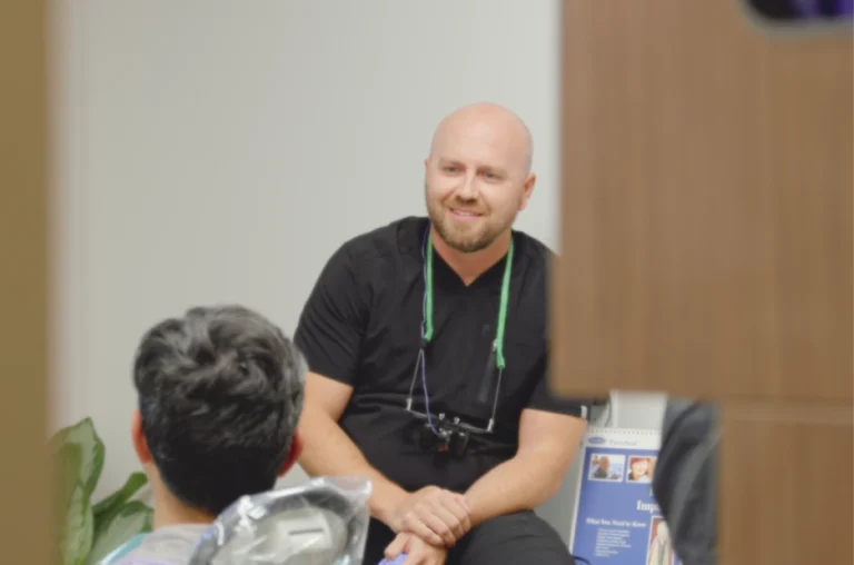 A dentist in black scrubs, with dental loupes around his neck, discusses the upcoming tooth extraction with a patient in the dental chair.